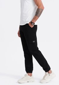 Černé cargo kalhoty se bočními kapsami, tapered střih, elastické manžety a strukturovaná látka. Kombinováno s bílým tričkem a světlými sneakers.