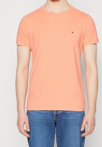 T-shirt a maniche corte in cotone corallo con colletto tondo e piccolo logo sul petto, abbinato a jeans in denim blu.