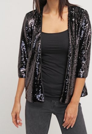 Frau trägt eine schwarze, mit Pailletten besetzte Jacke mit offenem Vorderteil, ein schwarzes ärmelloses Oberteil und dunkelgraue Jeans vor einem einfarbigen Hintergrund.