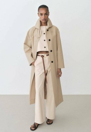 Cappotto lungo beige con colletto alto, bottoni neri, abbinato a un top beige corto e pantaloni beige a gamba larga, accentuato da una cintura marrone.