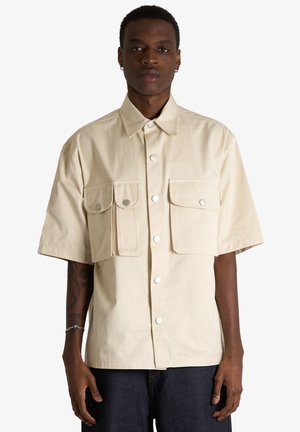 Giovane uomo che indossa una camicia beige a maniche corte con due grandi tasche frontali, in piedi davanti a uno sfondo semplice.