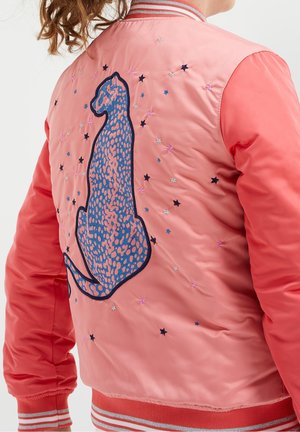 Personne portant une veste corail rose avec un patch léopard bleu et de petites étoiles brodées dans le dos.
