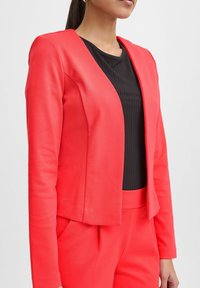 Figursydd korallfärgad blazer med långa ärmar, slät textur och öppen framdesign, kombinerad med en svart ribbad topp och matchande byxor.