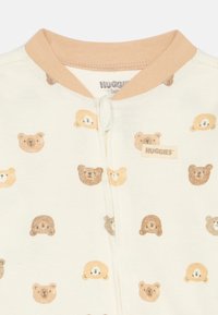 Body pentru bebeluși crem cu guler crem, închidere cu fermoar și un model repetat de fețe de urși maro, crem și bej. Eticheta "HUGGIES" pe piept.