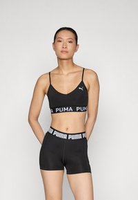 Crna sportska brusnica s tankim trakama i PUMA logotipom, uparena s crnim šortsevima koji imaju pojas s logotipom. Glatka tekstura, prilagođen dizajn.