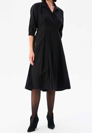 Femme portant une robe noire mi-longue à manches trois-quarts, des collants noirs transparents et des bottines noires à talons hauts.