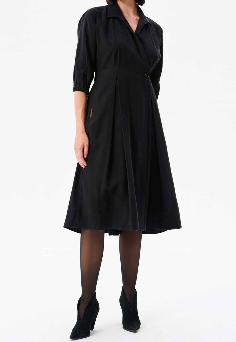 Femme portant une robe noire mi-longue à manches trois-quarts, des collants noirs transparents et des bottines noires à talons hauts.