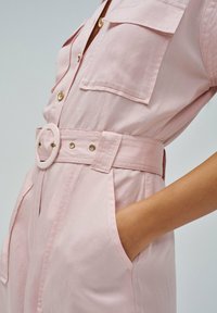 Vestido rosa claro con cinturón, grandes bolsillos frontales y botones dorados, con una mano colocada de manera casual en el bolsillo lateral.