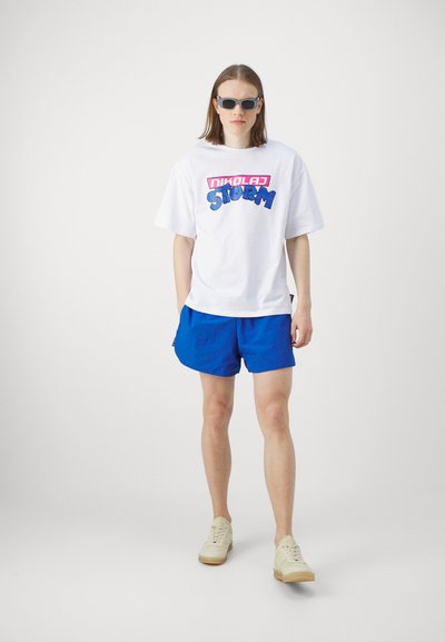 Valkoinen oversize T-paita, jossa on sininen ja vaaleanpunainen graafinen teksti edessä, yhdistettynä sinisiin shortseihin ja beigeihin sneakers-kengiin. Tummat aurinkolasit ovat päällä.