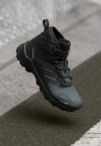 Schwarzer hochgeschlossener Wanderschuh mit strukturiertem Obermaterial, Netzakzenten und einer auffälligen Profilsohle. Verfügt über beschriftete Beschläge und ein Rautenmuster.