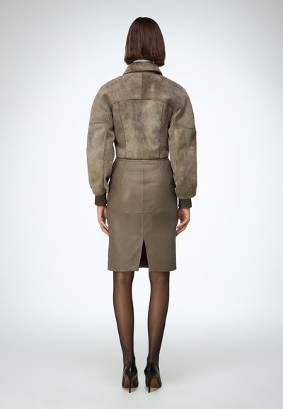 Smeđa bomber jakna od antilopa s rebrastim manžetama, uparena s kožnom suknjom maslinasto zelene boje do koljena s prorezom straga. Crne cipele na visoku petu.