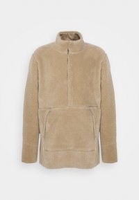 Beżowy polarowy sweter z wysokim kołnierzem, pół zamkiem błyskawicznym i kieszenią z przodu. Posiada miękki, textured materiał oraz długie rękawy.