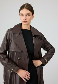 Bruine leren trenchcoat met een ingesneden kraag, dubbele knopen aan de voorkant en gouden kettingdetails op de mouwen. De textuur lijkt glad en glanzend.