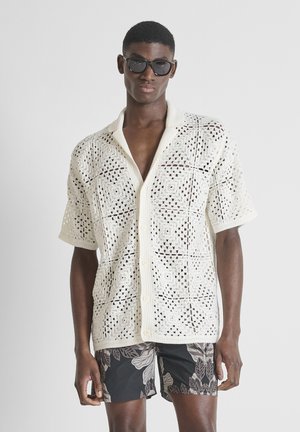 Hombre con gafas de sol negras, camisa blanca de crochet de manga corta con botones y pantalones cortos negros con estampado floral, de pie frente a un fondo sencillo.