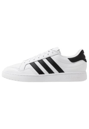 Baskets Adidas basses blanches avec trois bandes noires sur le côté, lacets blancs et languette de talon noire avec logo.