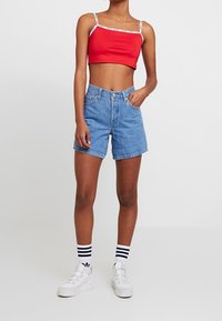 Kvinna som bär en röd kortärmad topp, blå denimshorts med hög midja, vita randiga strumpor och vita platåskor, stående med en hand i fickan.