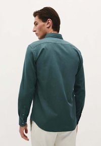 Camisetă lungă de culoare teal închis, cu guler cu nasturi, având o textură fină și manșete cu nasturi, văzută din spate.