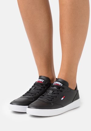 Schwarze Ledersneaker mit weißer Sohle, ausgestattet mit einem Schnürdesign, durchlöchertem Zehenbereich und einem Logo auf der Zunge in rot/weiß/blau.