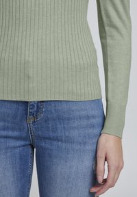Hellgrüner, gerippter Pullover mit langen Ärmeln, kombiniert mit blauen Jeans aus Denim, die leicht ausgewaschen sind und Distress-Details aufweisen.