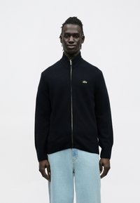 Camisola com fecho de correr azul-marinho com gola alta canelada, feita de tecido macio, com um pequeno logótipo verde no peito esquerdo e punhos canelados.