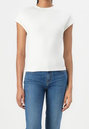 Personne portant un haut côtelé blanc ajusté à manches courtes et un jean en denim bleu, debout devant un fond clair uni.