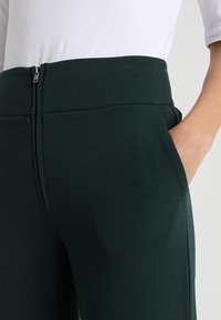 Mörkgröna högmidjade byxor gjorda av slät tyg, med en dold dragkedja och sidofickor. Enkel design med rena linjer.