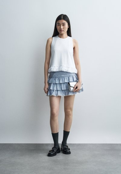 Νεαρή γυναίκα φοράει λευκή αμάνικη μπλούζα, μπλε φούστα με βολάν, μαύρα μοκασίνια με κάλτσες, κρατώντας ασημένιο φάκελο μπροστά σε απλό φόντο.