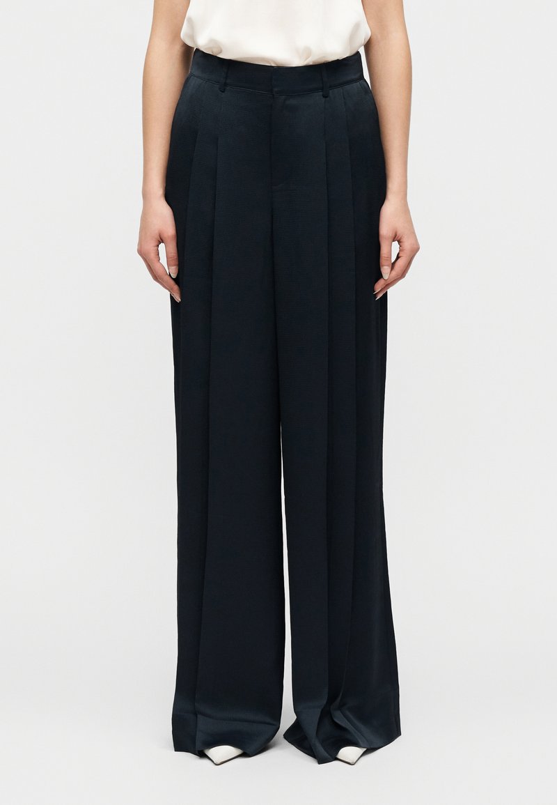 Γυναίκα που φοράει μαύρο παντελόνι με ψηλή μέση  και φαρδιά πόδια με πιέτες, συνδυασμένο με λευκά παπούτσια μυτερά σε απλό φόντο.