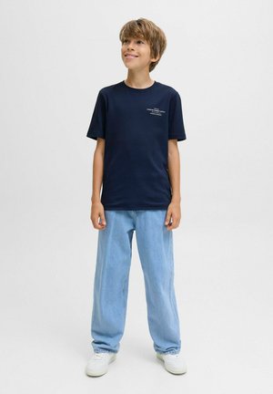 Ein lächelnder Junge steht in einem dunkelblauen T-Shirt und hellblauen Jeans, mit weißen Sneakers, vor einem schlichten weißen Hintergrund.