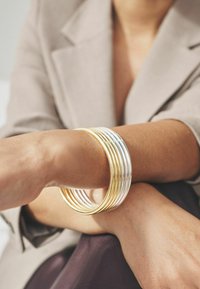 Mitmekordsed üksteise kohal asetsevad käevõrud kuldse ja hõbedase tooni, millel on sile ja poleeritud pind. Kandmine käel, millel on pehme ja tekstureeritud nahk.