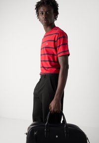 Rødstribet t-shirt med marineblå striber, korte ærmer. Sorte bukser, model holder en sort lædertaske med to håndtag og hardware detaljer.