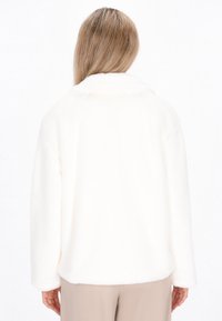 Person in beige Hosen, der eine weiße, flauschige Jacke mit langen Ärmeln trägt, von hinten vor einem weißen Hintergrund gezeigt.