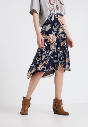 Femme portant une jupe midi fleurie bleu marine et des bottines marron, associée à un haut gris brodé, debout sur un fond blanc.