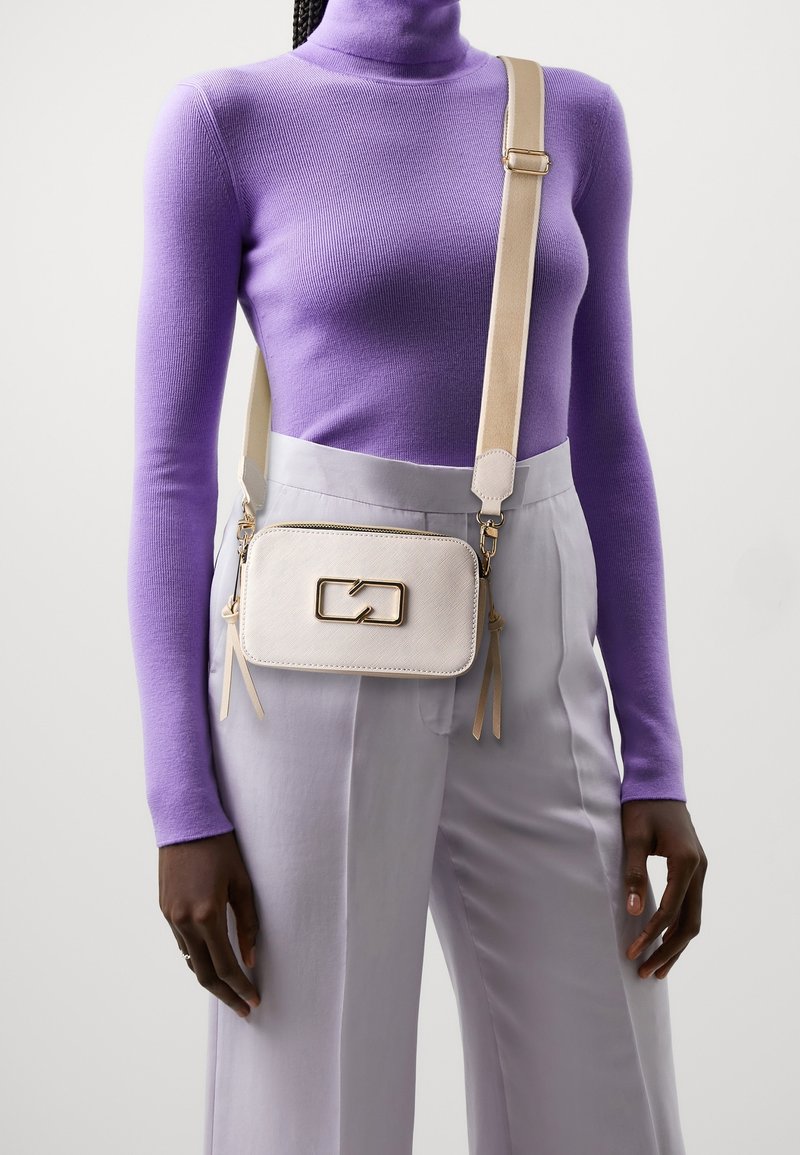 Personne portant un col roulé violet, un pantalon taille haute gris clair et un petit sac bandoulière crème avec une boucle dorée.