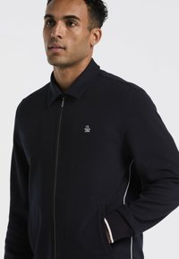 Marineblauwe ritsjack met een kraag, geribbeld textuur, zijzakken en witte accentstrepen. Bevat een klein logo op de borst.