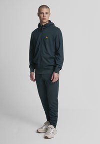 Dunkelgrüner Hoodie mit Reißverschluss und verstellbarem Kapuzen, kombiniert mit passenden Jogginghosen. Verfügt über ein kleines gelbes Logo auf der Brust.