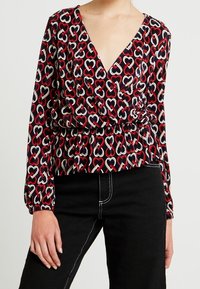 Blusa a maniche lunghe con scollo a V profondo, decorata con cuori rossi, bianchi e blu navy; vita arricciata e tessuto testurizzato.