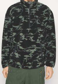 Schwarzer und grüner Fleecepullover mit hohem Kragen, halblangem Reißverschluss, Tarnmuster und zwei Reißverschlusstaschen vorne.