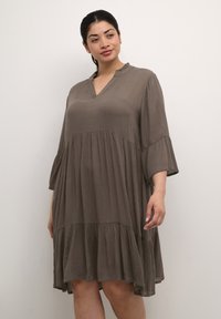 Robe sans manches à volants en tissu marron doux. Elle possède un décolleté en V et des accents froncés au col. Design fluide avec un ourlet droit.