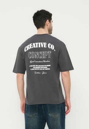 Férfi sötétszürke pólóban, amelyen fehér felirat olvasható: "CREATIVE CO. CONCEPT" és további szöveg a hátulján, kék farmerrel párosítva.