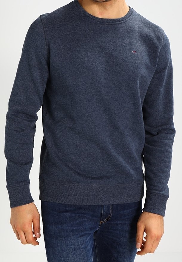Homme portant un sweat-shirt bleu foncé à col rond avec un petit logo sur la poitrine gauche, associé à un jean bleu, debout les bras détendus.