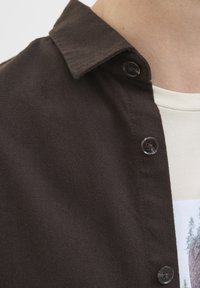 Primer plano de un cuello de camisa marrón oscuro con botones sobre una camiseta gráfica blanca que muestra árboles en un paisaje rocoso.