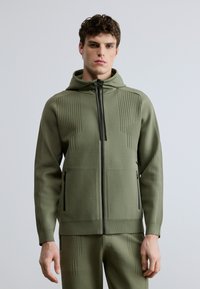 Chaqueta con capucha y cremallera en tela verde oliva, con patrones verticales acanalados, cremallera negra y dos bolsillos laterales con cierre de cremallera.