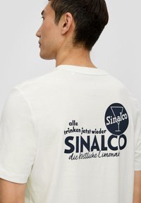 Valkoinen puuvillapaita, jossa on graafinen printti tummansinisellä värillä ja tekstillä "Sinalco" sekä cocktail-lasi -kuva takana.