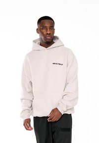 Sudadera oversized color beige claro con capucha ajustable, bolsillo frontal y logo "MULTIPLY" en negro en el pecho. Textura suave.