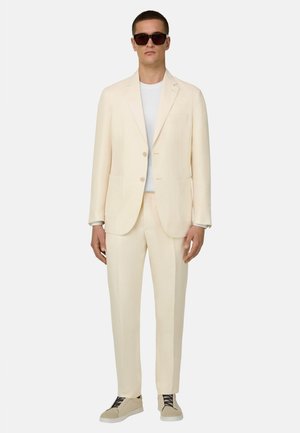 Homme portant un costume couleur crème avec un t-shirt blanc, des baskets beige et des lunettes de soleil foncées, debout devant un fond uni.