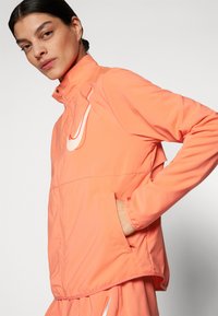 Veste légère zippée en corail vif, dotée d'un col haut, de poches latérales et d'un logo Nike blanc bien en évidence sur la poitrine.