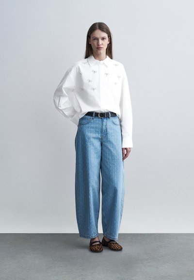 Γυναίκα που φοράει άσπρο πουκάμισο με κουμπιά και μικρά σχέδια φιόγκων, φαρδιά ανοιχτόχρωμα μπλε τζιν, μαύρη ζώνη και φλατ παπούτσια με λεοπάρ σχέδιο, στέκεται σε γκρι πάτωμα.