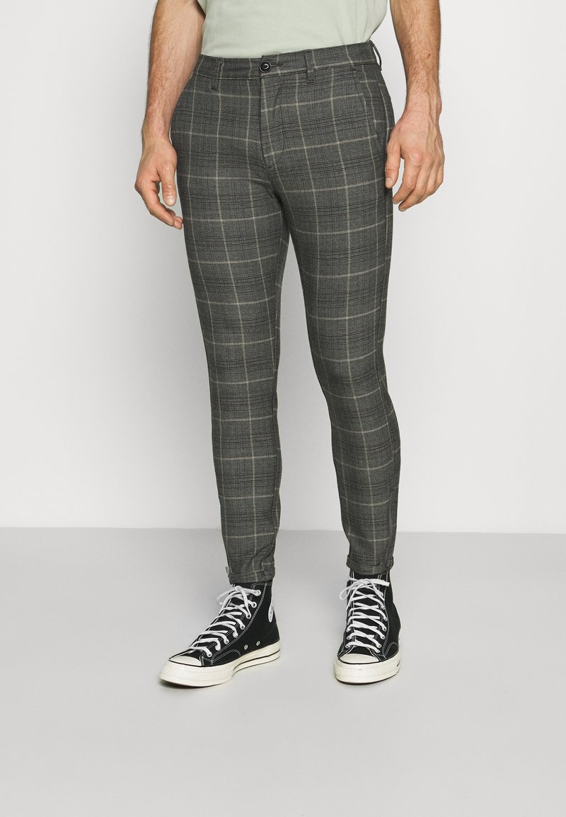 Man som bär slim-fit grå rutiga byxor och svarta högklackade sneakers med vita snören, stående mot en neutral bakgrund.