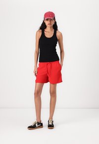Zwarte mouwloze tanktop, rode shorts met elastische band, gecombineerd met een roze pet en zwarte sneakers met tan accenten.